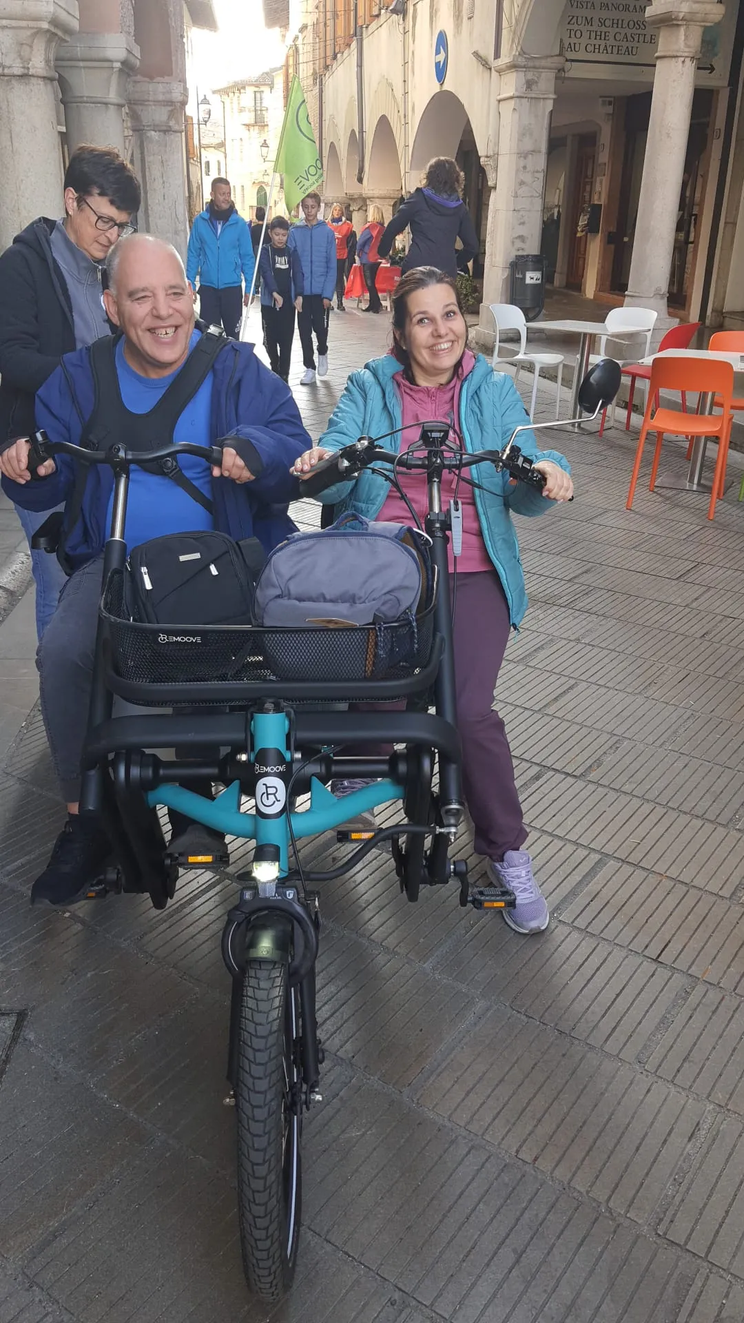 Una donna accompagna in bici un uomo su una special bike sotto i portici di un centro storico, tra i tavolini di un bar.