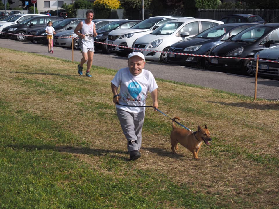 Un uomo con maglietta Willeasy corre con un cane al guinzaglio durante una gara podistica, su un prato accanto a un parcheggio