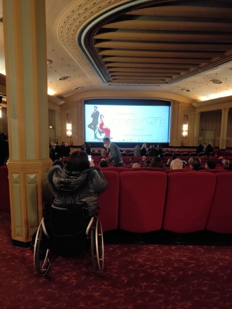 Una donna su una carrozzina assiste alla proiezione del film Corro da te in una sala cinematografica, tra il pubblico seduto