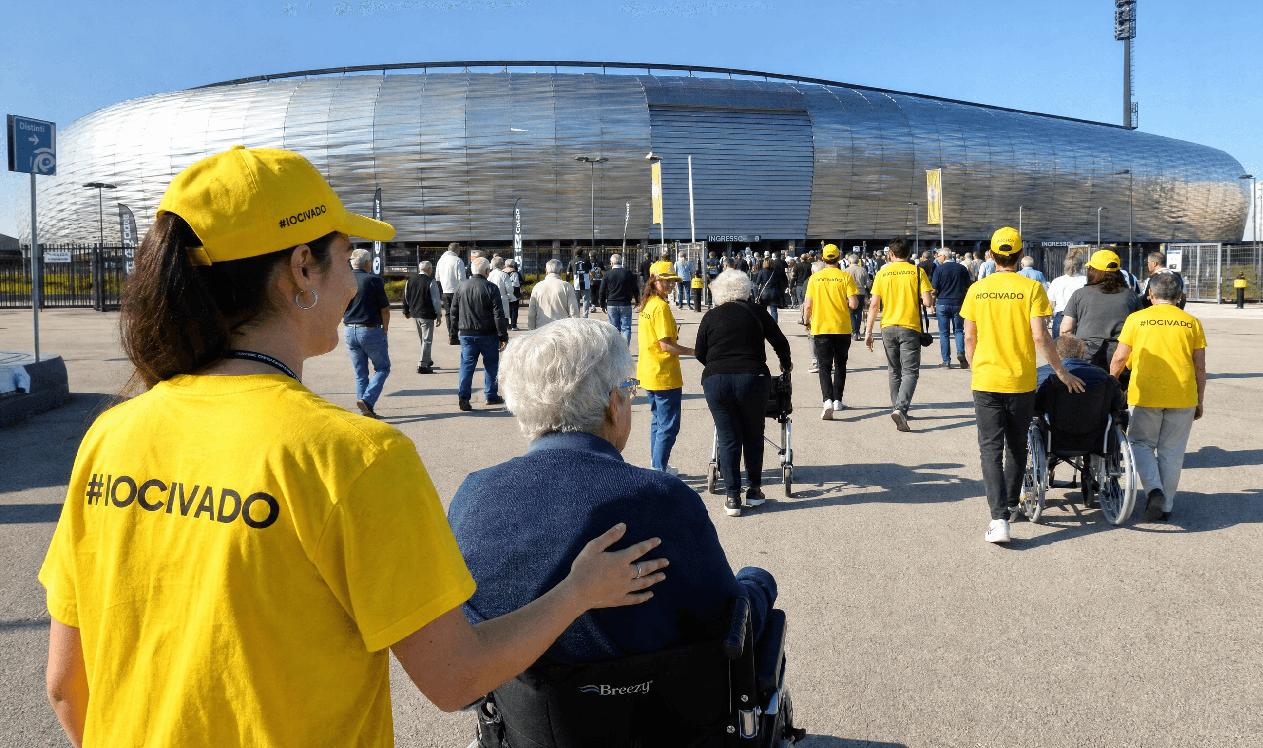 Una volontaria IO CI VADO con maglietta gialla "#IOCIVADO" accompagna una signora in sedia a rotelle verso l'ingresso della Dacia Arena, tra altri volontari e tifosi in fila per entrare.